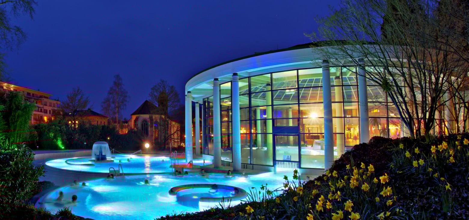 Abendaufnahme der Caracalla Therme in Baden-Baden mit beleuchtetem Außenbecken und moderner Glasfassade, umgeben von blühenden Narzissen im Vordergrund.