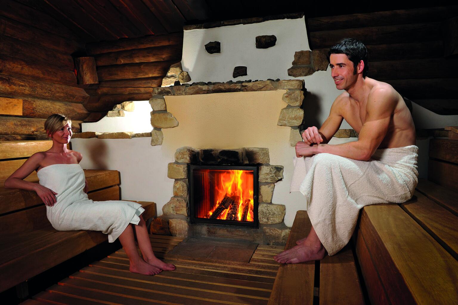 Zwei Personen sitzen in Handtüchern in einer Holzsauna und blicken auf einen Kamin mit brennendem Feuer.
