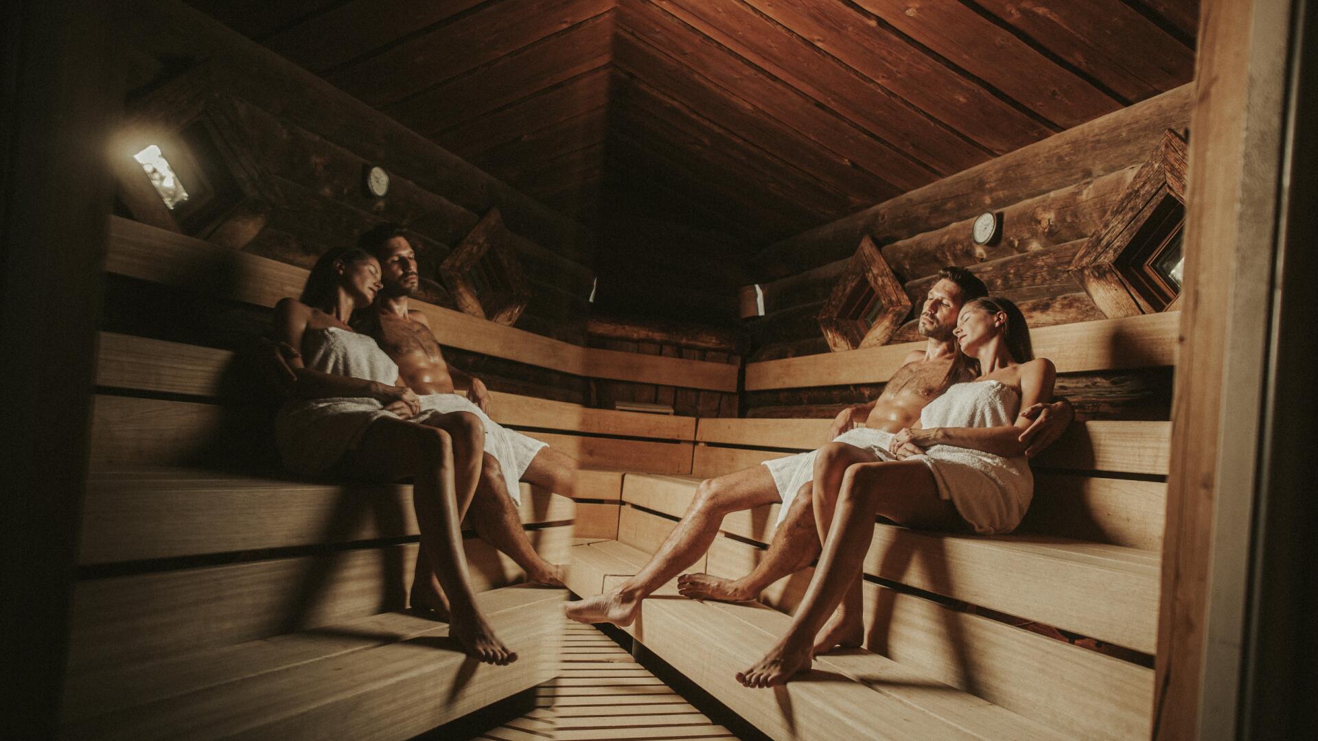 Innenansicht einer Waldsauna mit rustikaler Holzverkleidung. Zwei Paare sitzen entspannt in Handtücher gehüllt auf breiten Holzbänken und genießen die Wärme. Gedämpftes Licht und natürliche Materialien schaffen eine gemütliche, naturnahe Atmosphäre für erholsame Saunagänge.