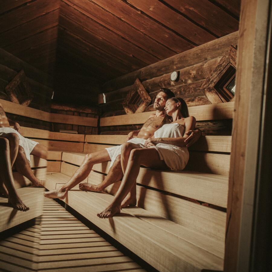Innenansicht einer Waldsauna mit rustikaler Holzverkleidung. Zwei Paare sitzen entspannt in Handtücher gehüllt auf breiten Holzbänken und genießen die Wärme. Gedämpftes Licht und natürliche Materialien schaffen eine gemütliche, naturnahe Atmosphäre für erholsame Saunagänge.