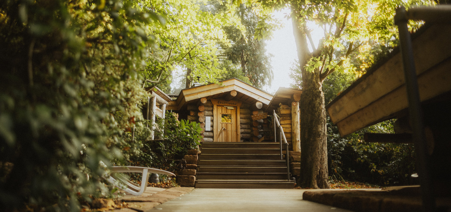 Blockhaus-Sauna im Schlossgarten der Caracalla Therme Baden-Baden. Der Holzsteg führt durch den grünen Schlossgarten mit hohen Bäumen und dichter Vegetation direkt zum Eingang des kleinen Saunahauses aus massiven Holzstämmen. Die Umgebung wirkt idyllisch und naturnah.